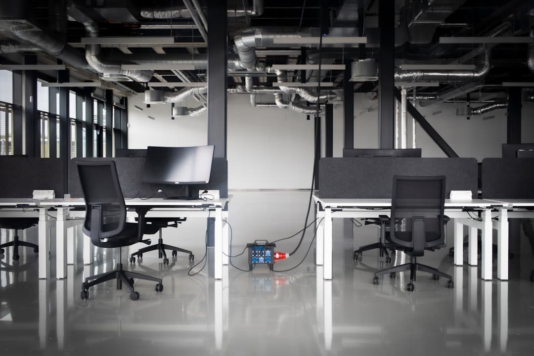 Empty modern open-plan office with rows of vacant desks and computer monitors, natural light from large windows.